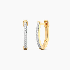 A close-up of a woman’s ear showcases the ELLA STEIN - SLIM PAVE DIAMOND HUGGIE EARRINGS (MEDIUM) by ELLA STEIN, gold plated with sparkling pave diamonds. Her hand, wearing a simple gold ring, gently touches her loose, wavy blonde hair.