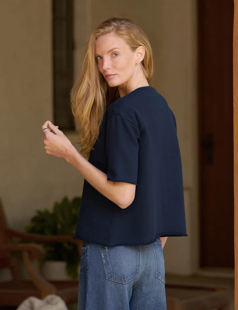 A woman with long blonde hair stands indoors with her hands in her pockets, looking at the camera. She is wearing blue raw hem jeans and the FRANK & EILEEN Perry Sweatshirt Tee in British Royal Navy from the California Heritage Collection. A wooden post and a green plant are visible in the background.