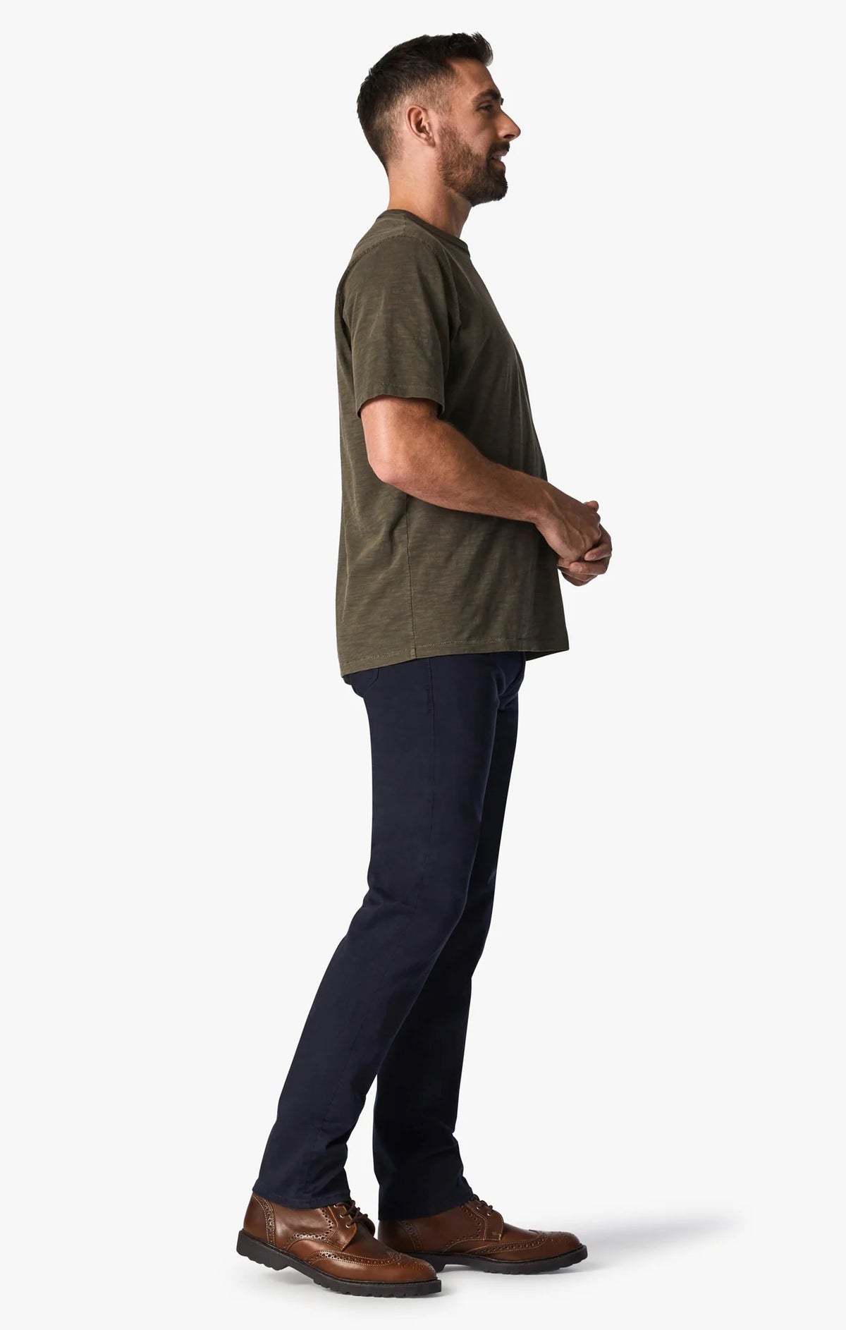 A man with short hair and a beard stands facing forward, wearing an olive green T-shirt, 34 HERITAGE - COURAGE STRAIGHT LEG PANT IN NAVY TWILL, and brown shoes against a plain white background.