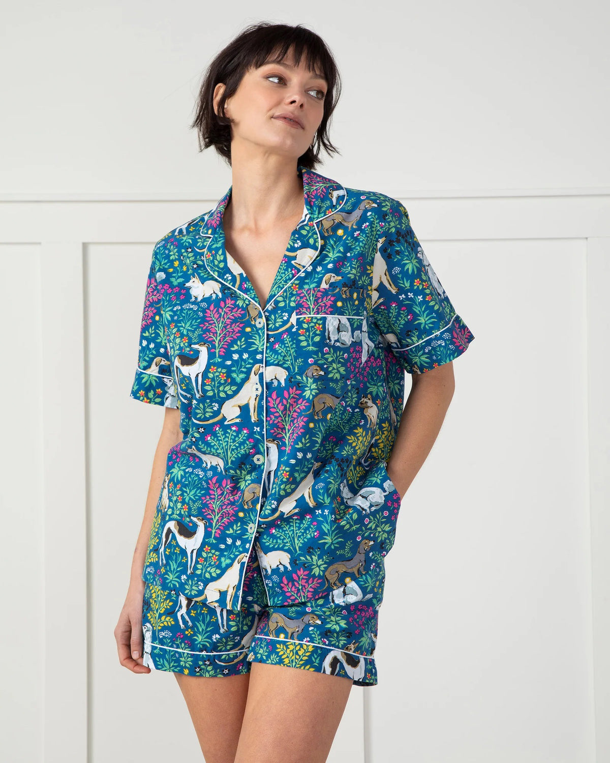 A person is wearing the PRINTFRESH &quot;Must Love Dogs&quot; pajama short set in organic cotton as they stand barefoot on a woven rug against white paneled walls, facing away from the camera.