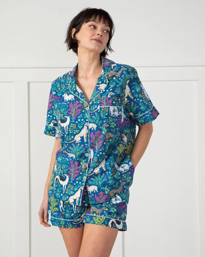 A person is wearing the PRINTFRESH "Must Love Dogs" pajama short set in organic cotton as they stand barefoot on a woven rug against white paneled walls, facing away from the camera.