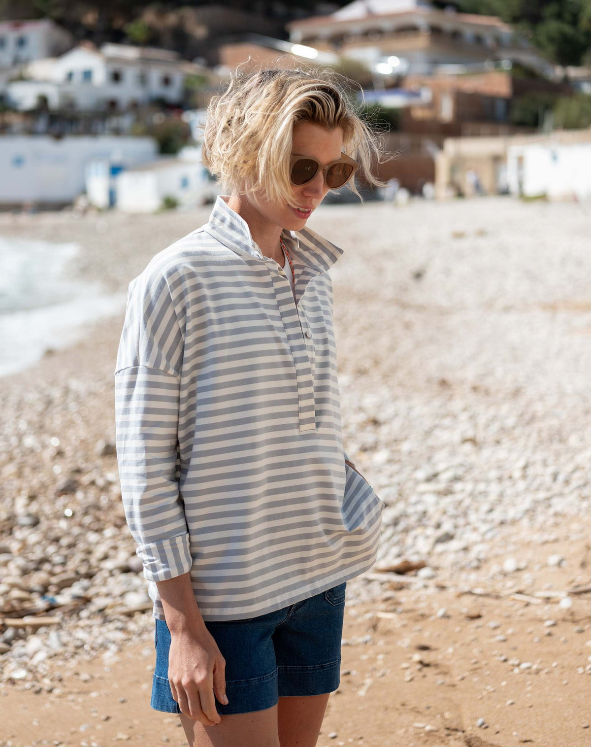 A person wearing sunglasses, denim shorts, and the MERSEA Emerson Pop Up Collar Top in Slate Blue Stripe stands on a rocky beach with buildings and trees in the background.