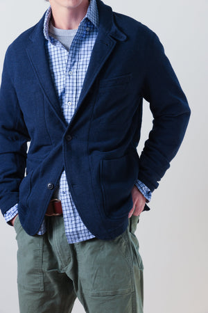 A person wearing the RELWEN - PIQUE TRAP BLAZER IN NAVY HEATHER over a checked shirt and green cargo pants stands with hands in pockets against a plain background, showcasing the blazer’s relaxed fit.