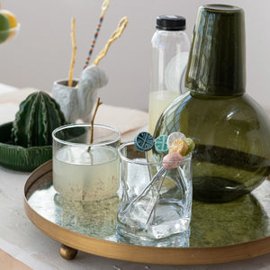 A round tray displays two CREATIVE COOP HAND-BLOWN ROUND CONFETTI DRINKING GLASSES with beverages, decorative stirrers, a green bottle, lemonade bottle, and a small cactus ornament on a tabletop with light linens.