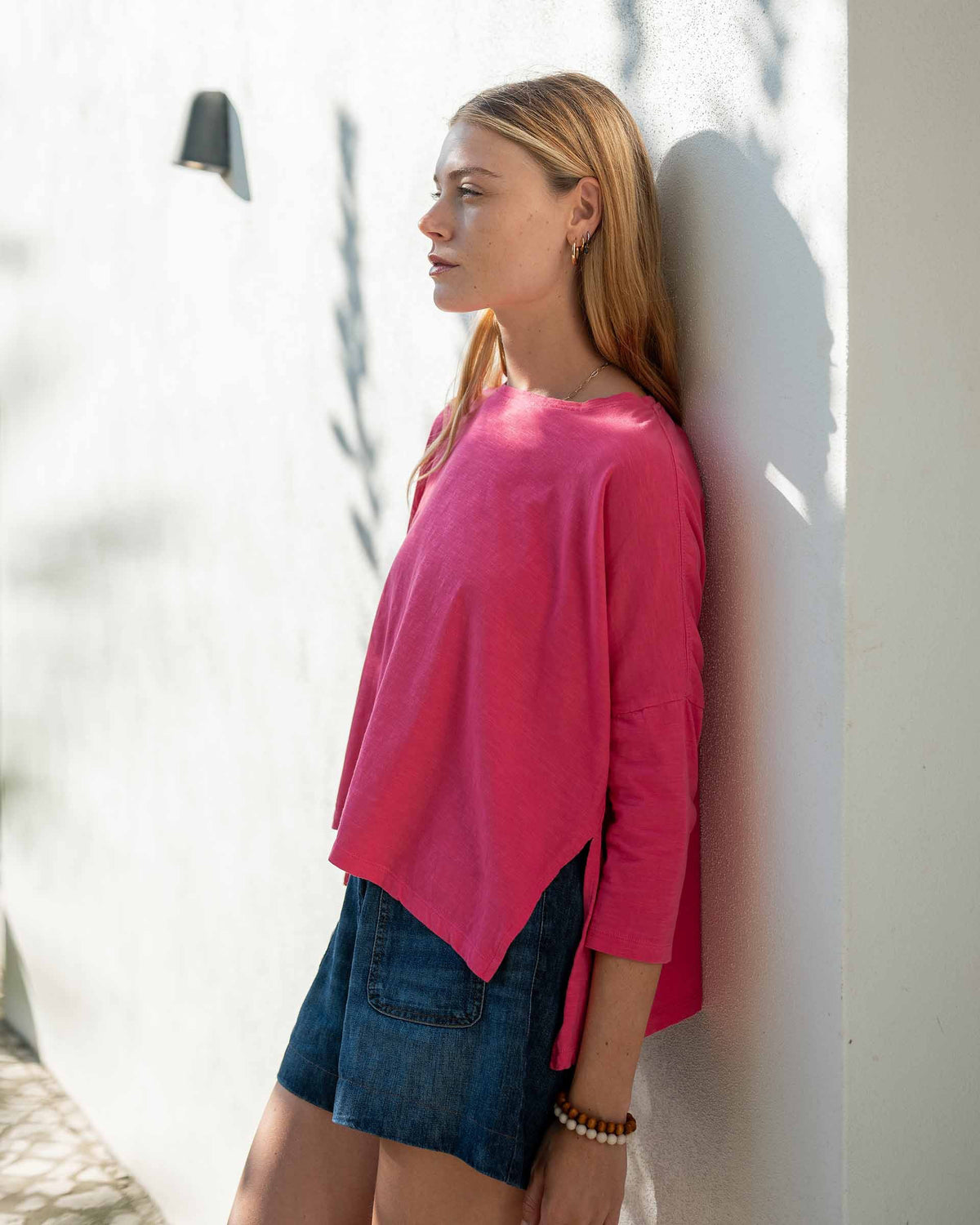 A woman with long, straight hair stands against a white wall, wearing the MERSEA Catalina Slub Tee in blue paired with white pants. The midweight textured cotton tee is from MERSEA. She looks at the camera with a neutral expression.