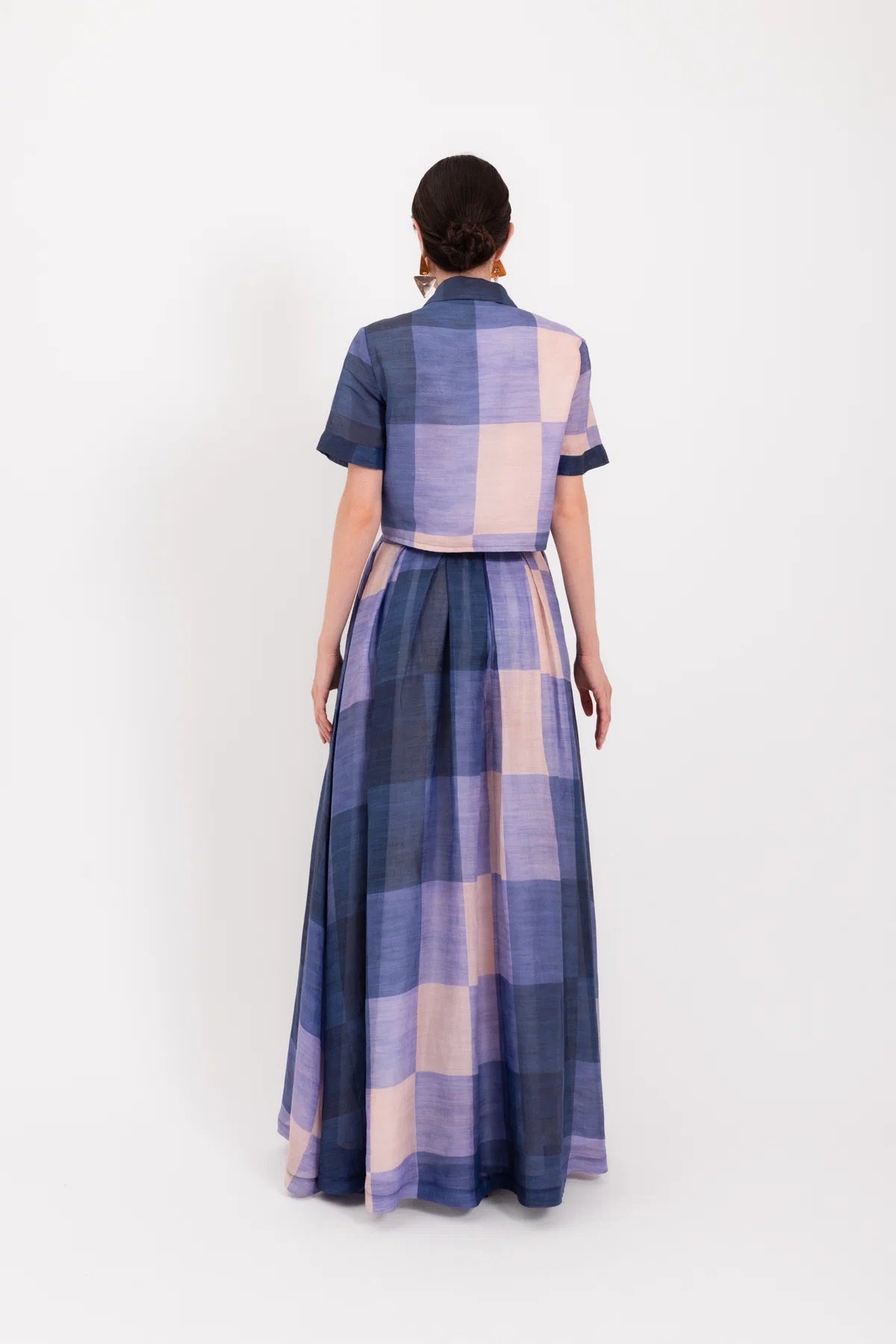 A woman wearing oversized orange sunglasses and the LAVI - MENTA Maxi Skirt in Quadri by LAVI, a long blue and beige plaid skirt with a tied waist and flowing maxi silhouette, stands against a plain white background.