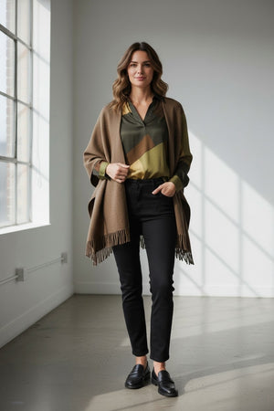 A woman stands indoors by a large window, sunlight streaming in and casting soft shadows on the floor, wearing the HARTFORD - VIRGINIE WOOL CAPE — a chic and casual brown cape from HARTFORD crafted from double-faced wool — layered over a patterned blouse, black pants, and black loafers.