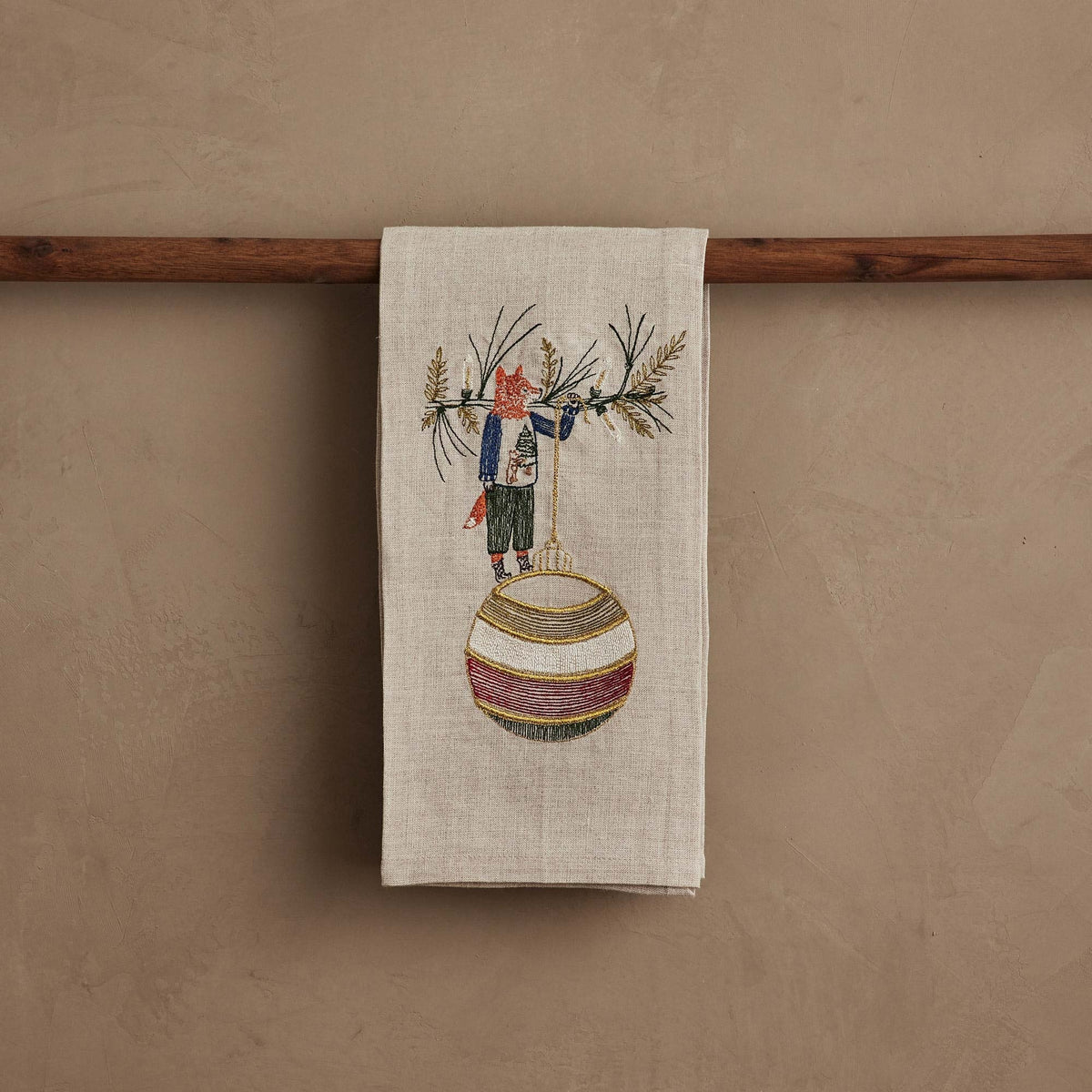 A beige linen DECK THE HALLS FOX LINEN TEA TOWEL embroidered with a fox holding a large, striped Christmas ornament, perfect for vintage Christmas kitchen decor.