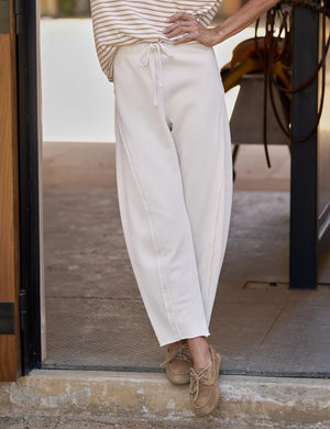 A person stands in a doorway wearing the FRANK & EILEEN Bibi Barrel Leg Sweatpant in Triple Fleece Vintage White, featuring raw edge side seams. Only the lower half of their body is visible. They pair the sweatpants with a striped top and tan boat shoes, with one hand resting on their hip.