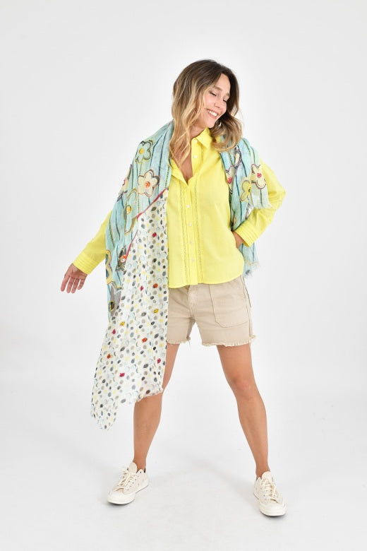 A woman in a yellow sweater and beige skirt holds up the STORIATIPIC - ANNE SCARF IN AQUA, a colorful linen scarf by STORIATIPIC featuring floral and polka dot patterns with exquisite handmade embroidery, against a white background.
