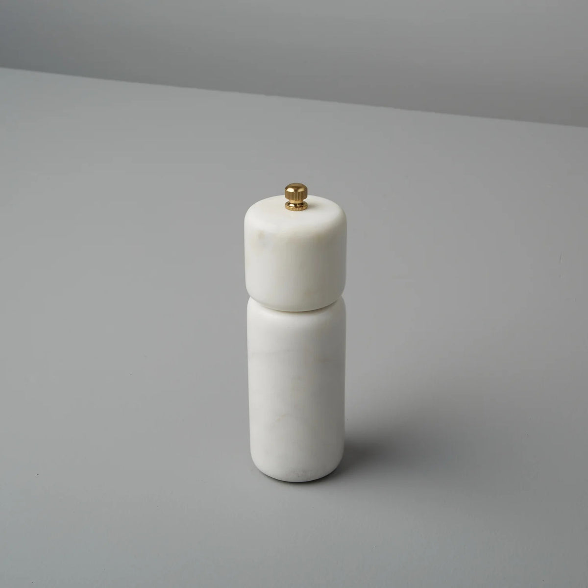 The MARBLE PEPPER GRINDER by BE HOME, INC is a minimalist white marble grinder, ethically crafted to showcase the natural beauty of stone. It features a sleek cylindrical shape topped with a small gold knob and stands elegantly upright on a plain gray surface.