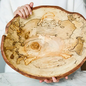 The SPENCER PETERMAN - SPALTED OVAL LIVE EDGE BOWL 15" by PETERMANS BOARDS & BOWLS INC, crafted from upcycled spalted maple wood and featuring a natural uneven rim, rests on a light gray marble surface against a plain white background.