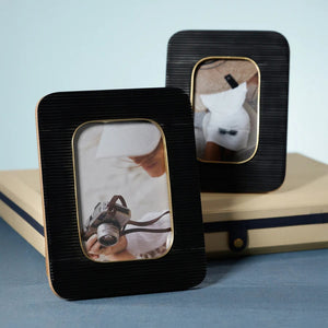 Two ZODAX BLACK RESIN RIBBED PHOTO FRAMES WITH BRASS ACCENT (5x7) display photos of a person in white, one with a camera. Each frame rests on beige boxes against a light blue background.