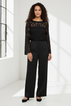A woman with shoulder-length dark hair is wearing the HINSON WU - BLAKELY LACE TOP, a long-sleeve black lace top that features flared sleeves, paired with black pants. She stands against a plain light gray background, neutrally expressing herself with one hand on her hip.