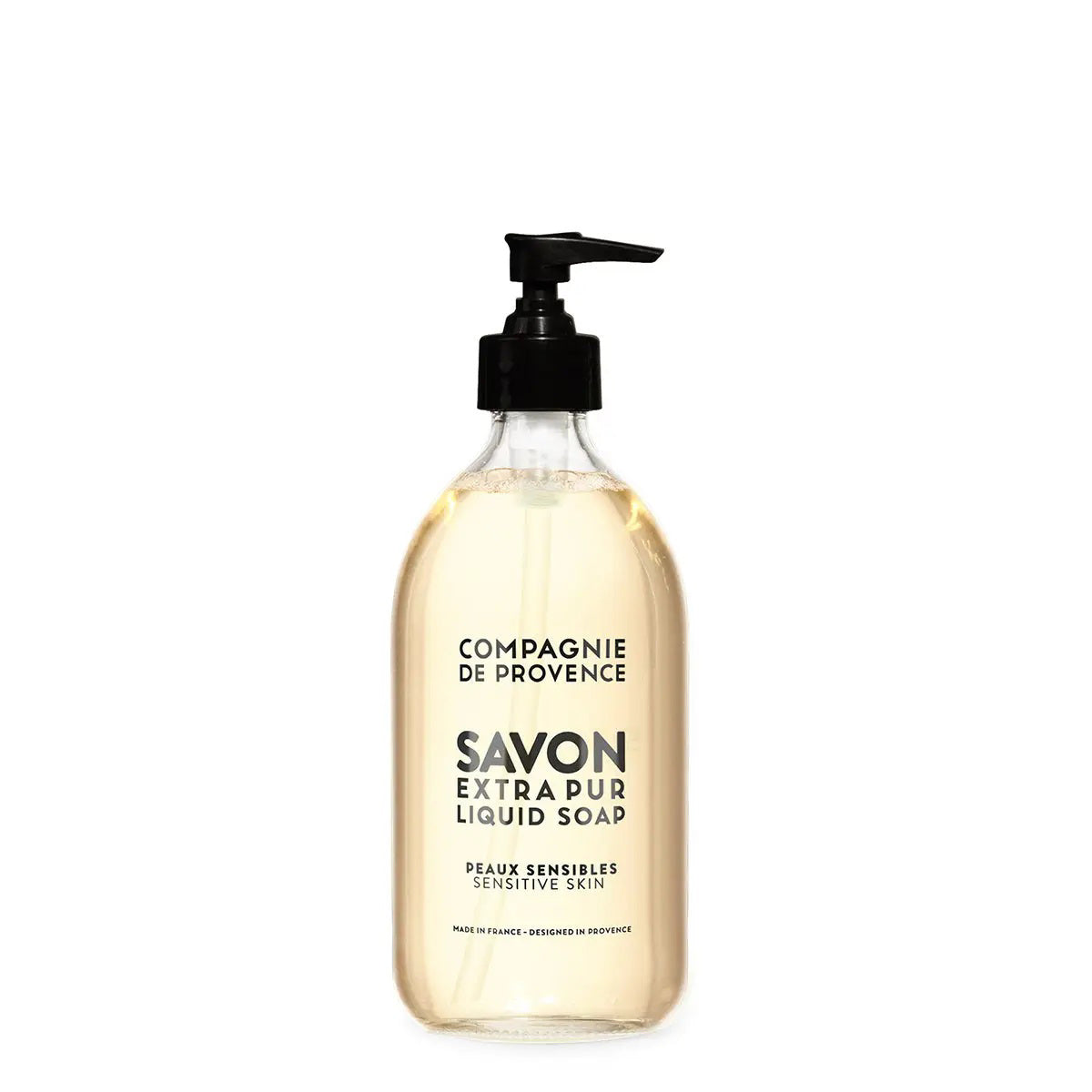A bottle of CIE LUXE BRANDS COMPAGNIE DE PROVENCE SAVON EXTRA PUR LIQUIDE SENSITIVE SKIN 10OZ is displayed next to a white oval stone, green grapes, a white flower, and green foliage on a beige surface with a neutral background.