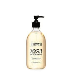 A bottle of CIE LUXE BRANDS COMPAGNIE DE PROVENCE SAVON EXTRA PUR LIQUIDE SENSITIVE SKIN 10OZ is displayed next to a white oval stone, green grapes, a white flower, and green foliage on a beige surface with a neutral background.