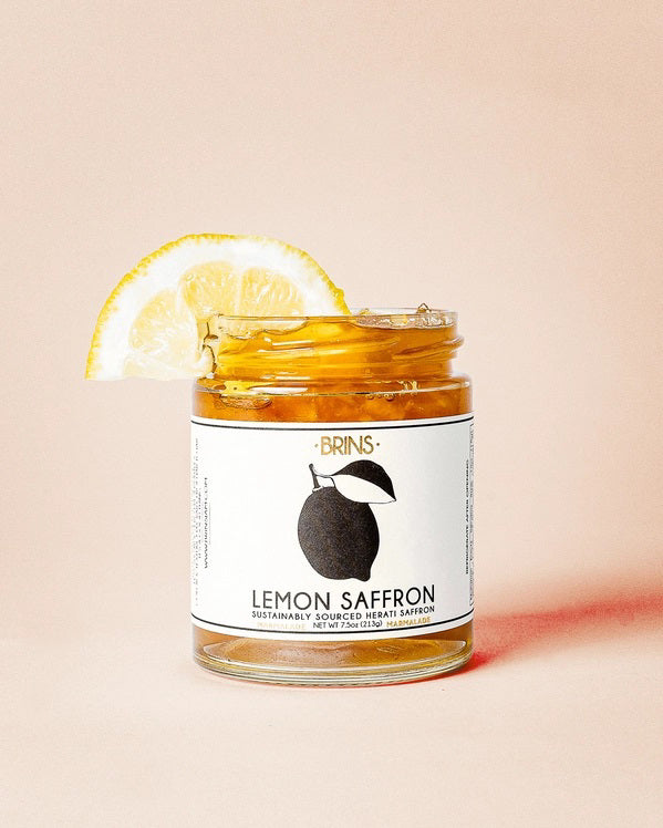 A jar labeled “BRINS - LEMON SAFFRON MARMALADE” from BRINS, garnished with a lemon slice, sits against a light beige background. The jar holds a vibrant yellow marmalade crafted from whole fresh lemons and infused with aromatic saffron for a bright, floral flavor.