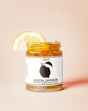 A jar labeled “BRINS - LEMON SAFFRON MARMALADE” from BRINS, garnished with a lemon slice, sits against a light beige background. The jar holds a vibrant yellow marmalade crafted from whole fresh lemons and infused with aromatic saffron for a bright, floral flavor.