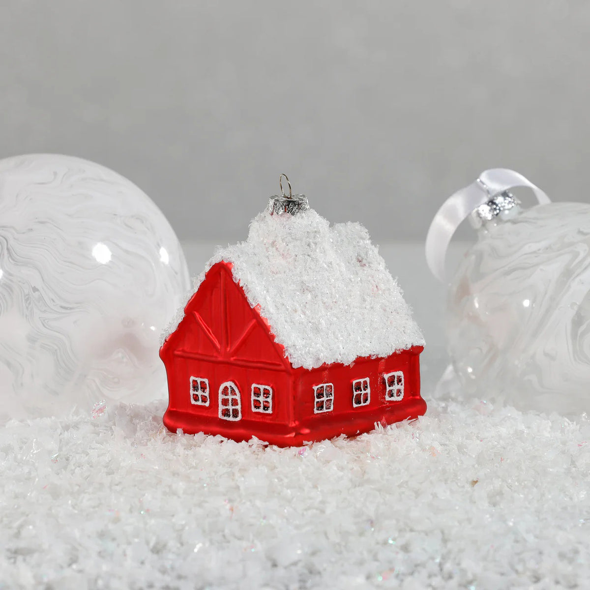 The ZODAX GLASS RED HOUSE ORNAMENT features a snow-covered roof and sits on faux snow, flanked by two clear glass baubles with white swirls—an ideal size in both inches and centimeters for festive displays.