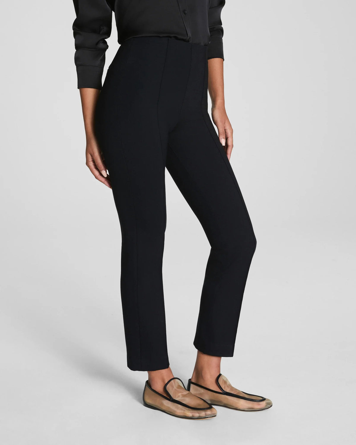 A person wears SPANX&#39;s Ponte Ankle Slim Pant in Black with a black long-sleeve top and beige loafers. Only their lower half is visible, standing against a plain, light background.