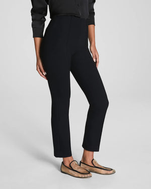 A person wears SPANX's Ponte Ankle Slim Pant in Black with a black long-sleeve top and beige loafers. Only their lower half is visible, standing against a plain, light background.
