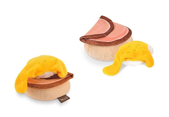Introducing the PLAY PET LIFESTYLES/BRANDS BARKING BRUNCH - BENNY’S BENEDICT: an adorable dog toy designed to look like a plush egg with a yellow yolk atop a round, brown base, mimicking a classic brunch favorite. Perfect for playful pups, this fun and charming brunch-themed toy features a black fabric tag attached at the bottom.