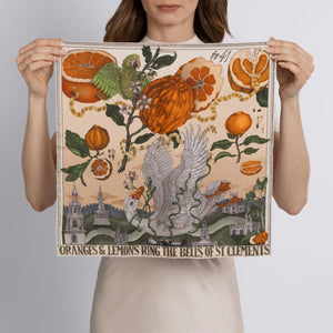 A woman in a beige short-sleeved top and matching skirt stands against a light background, adorned with the SABINA SAVAGE ORANGES AND LEMONS SILK TWILL SCARF in Cintrine/Zest. Her shoulder-length brown hair softly frames her face, enhancing her effortless elegance.