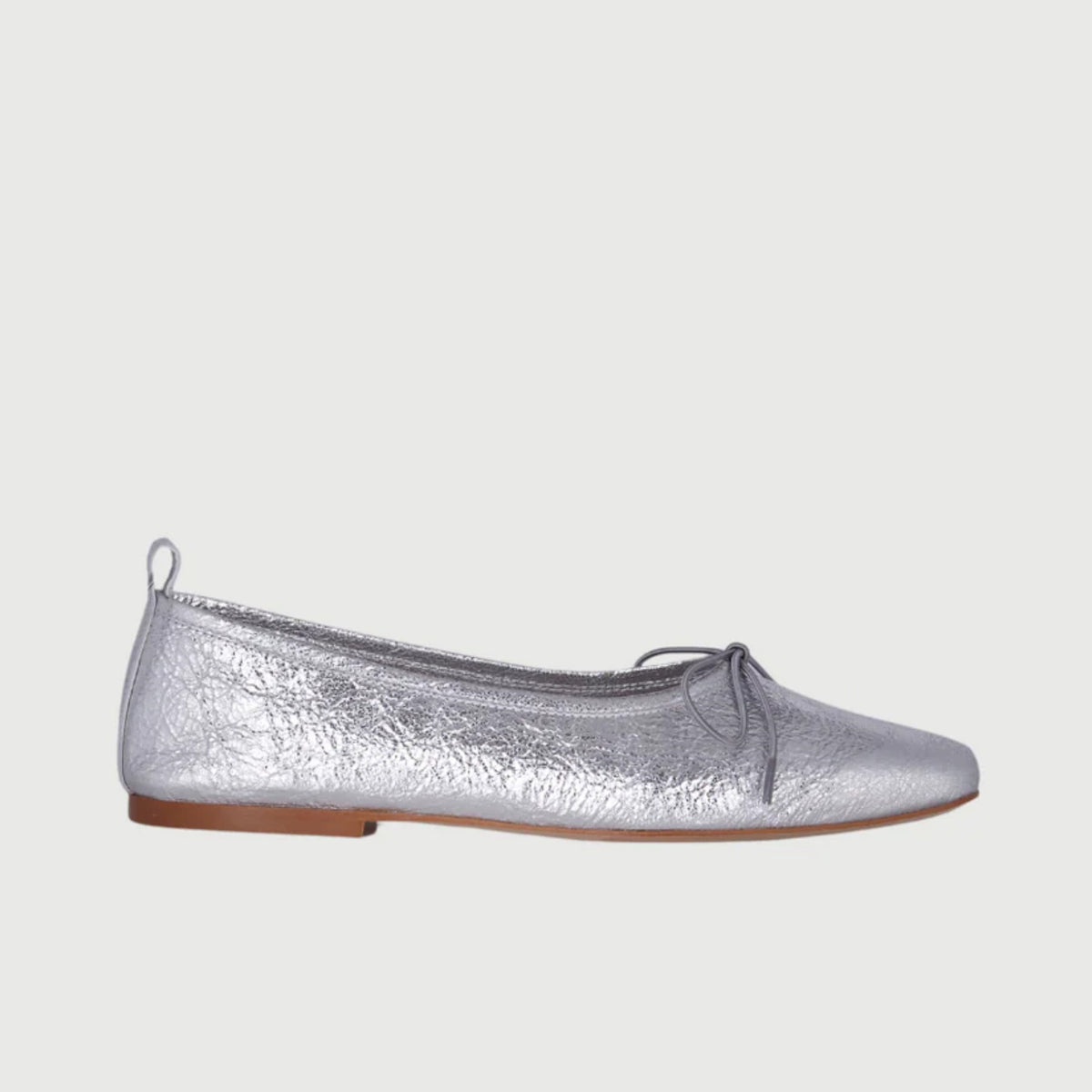 The Augusta - Sienna Ballet Flat by AUGUSTA is a single silver-colored shoe featuring a subtle metallic sheen and a simple bow on the front. It has a low brown sole and a pull tab at the back. Handcrafted in Spain from leather, this ballet flat is shown from the side against a light gray background.