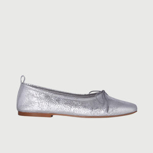 The Augusta - Sienna Ballet Flat by AUGUSTA is a single silver-colored shoe featuring a subtle metallic sheen and a simple bow on the front. It has a low brown sole and a pull tab at the back. Handcrafted in Spain from leather, this ballet flat is shown from the side against a light gray background.