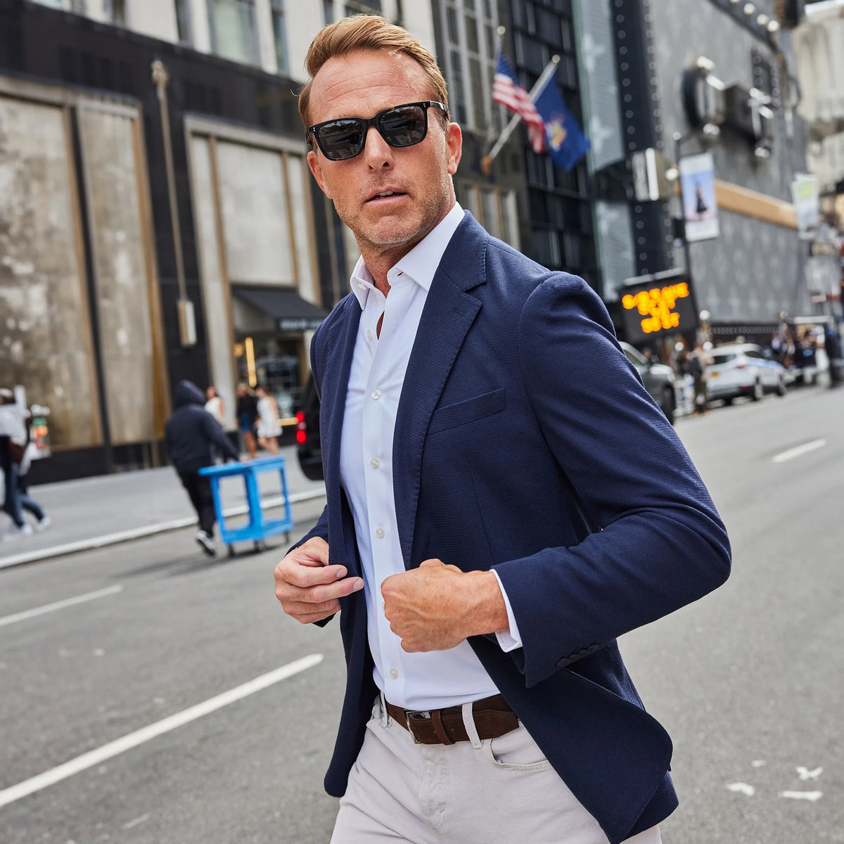 A man in the COLLARS AND CO Maverick Knit Unstructured Blazer in navy, paired with a light blue shirt and sunglasses, stands on a busy city street with cars, bikes, and buildings behind him.