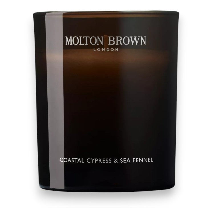 A dark glass MOLTON BROWN - COASTAL CYPRESS 1 WICK CANDLE, labeled &quot;Molton Brown London&quot; with &quot;Coastal Cypress &amp; Sea Fennel&quot; beneath, photographed on a white background, evokes marine notes and the freshness of sea fennel.