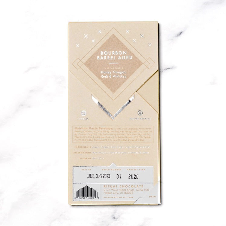 The image shows a light brown rectangular package of Ritual Chocolate labeled &quot;RITUAL CHOCOLATE - BOURBON BARREL AGED CHOCOLATE BAR 75%.&quot; This limited edition chocolate, crafted from aged cacao nibs, features geometric designs in silver and white and contains 2.12 oz (60g) of chocolate. The background is a marble surface.