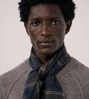 A man wearing a grey long-sleeve sweater and the HARTFORD - TARTAN WOOL SCARF IN INDIGO, GREY AND CAMEL stands against a white background, looking directly at the camera with his hands in his pockets—showcasing this classic men’s winter accessory from HARTFORD.