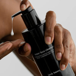 A person with short curly hair gently massages JACK HENRY - SUPER FACE CLEANSER onto their face, while ingredient callouts highlight bamboo powder for exfoliation, kakadu plum for brightening, and willow bark for cell renewal.