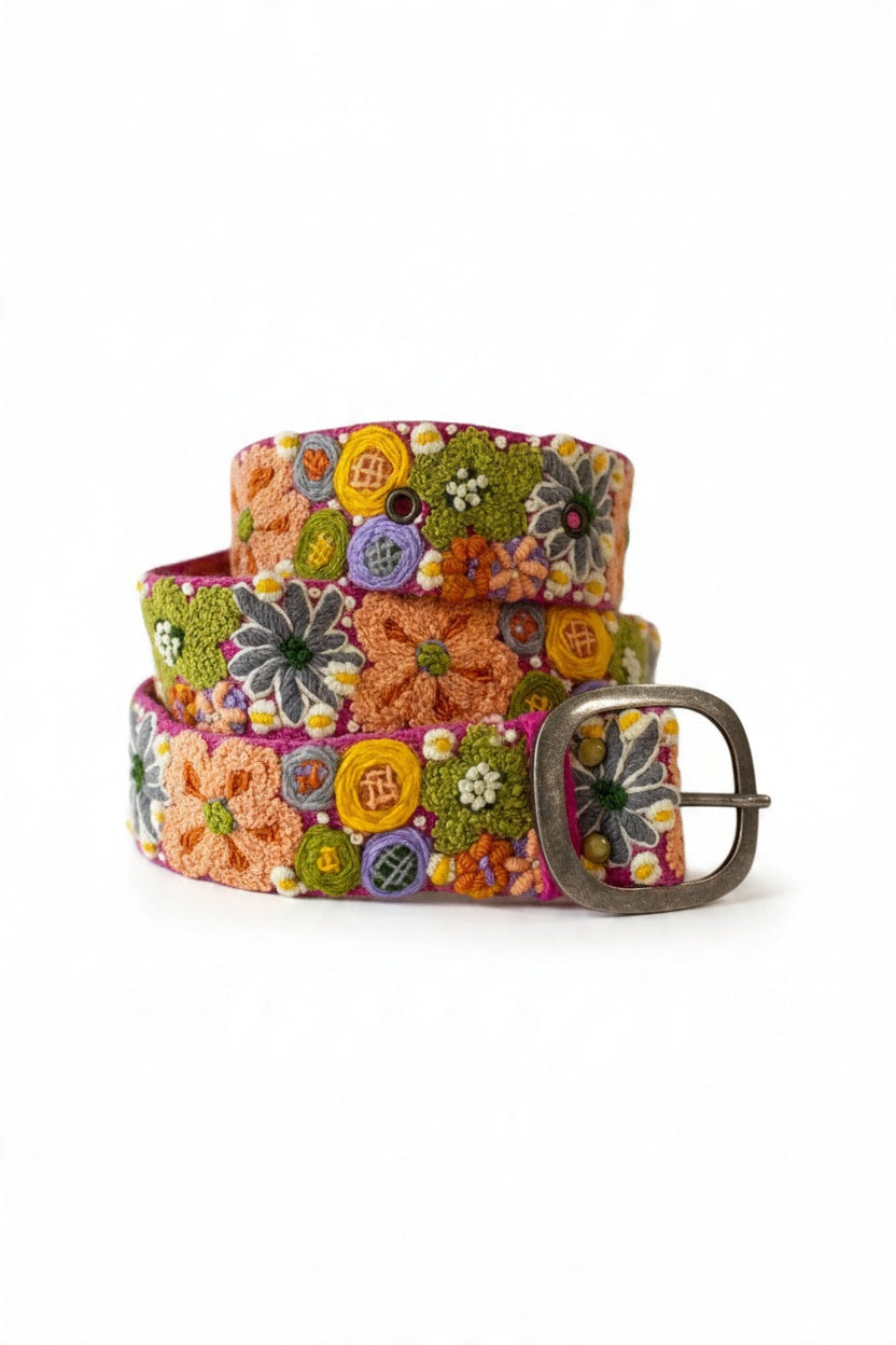 The JENNY KRAUSS WILDFLOWERS EMBROIDERED WOOL BELT features hand-embroidered floral patterns in orange, yellow, green, purple, and white on a peach background, crafted in Peru with a silver buckle and shown coiled on a white surface.