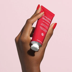 With perfectly manicured nails, a person applies MANUCURIST - COSMOS ORGANIC HAND CREAM, ROSE NOUVELLE from MANUCURIST INC to the back of their hand against a soft pink background, emphasizing the importance of caring for hands and nails.