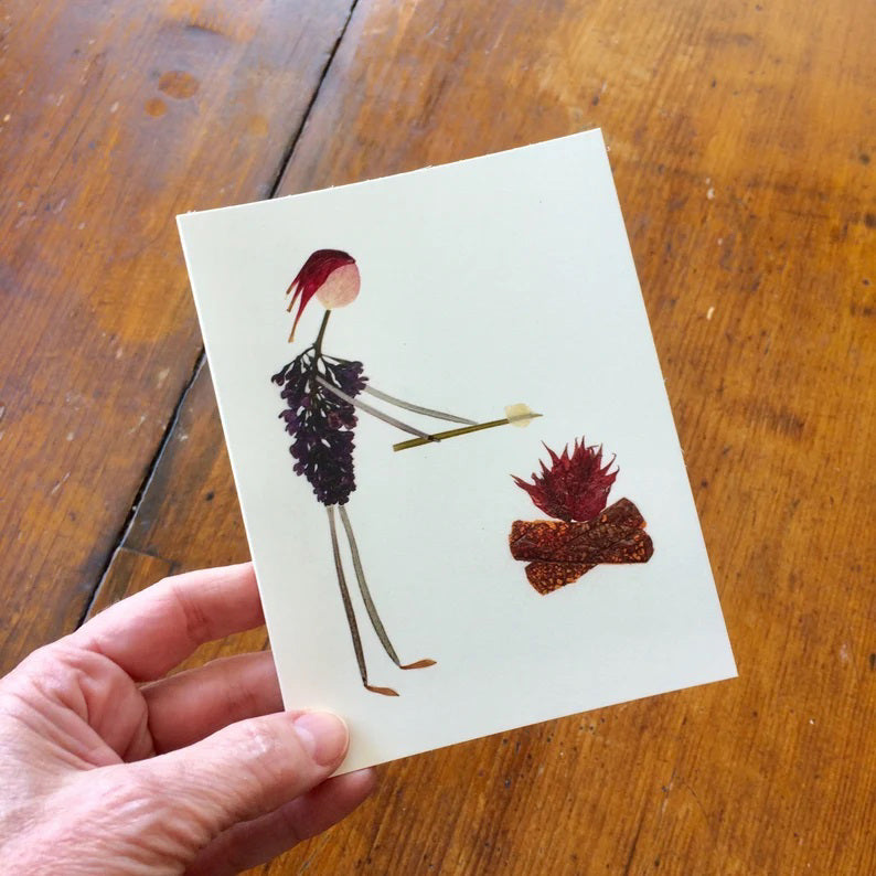 The PETAL PEOPLE PRESS - CAMP FIRE CARD by PETAL PEOPLE PRESS is a charming greeting card showcasing a stick figure made from dried flowers and leaves, roasting marshmallows. The design features purple flowers as the body and red petals with brown leaves for the campfire on a white background.
