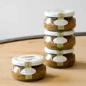 A jar of BELLA CUCINA - PORCINI & PARMESAN PESTO is displayed against a light gray background. The jar is topped with a white lid and adorned with a green label featuring an illustration of porcini mushrooms, alongside oval text labels indicating the product name and net weight of 6 oz (168 g).
