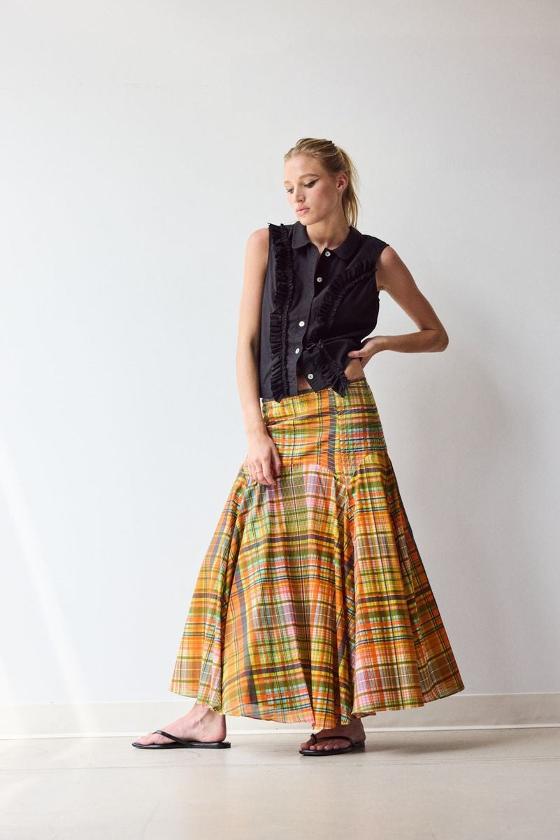 A woman stands indoors against a plain wall, wearing the HOPE FOR FLOWERS Circle Skirt in Spring Plaid, crafted from GOTS certified organic cotton lawn. She pairs it with a sleeveless black button-up top, black sandals, and has her hair tied back.