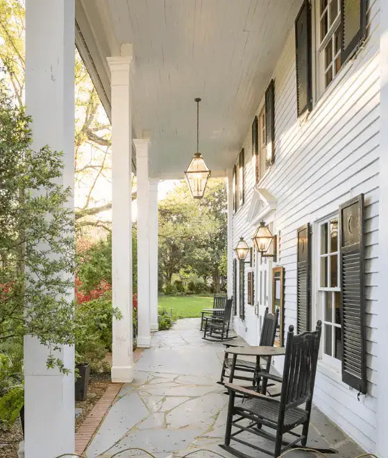 A wide porch of a white house with black shutters, featuring hanging lanterns, rocking chairs, and stone flooring, overlooking a green lawn and garden.