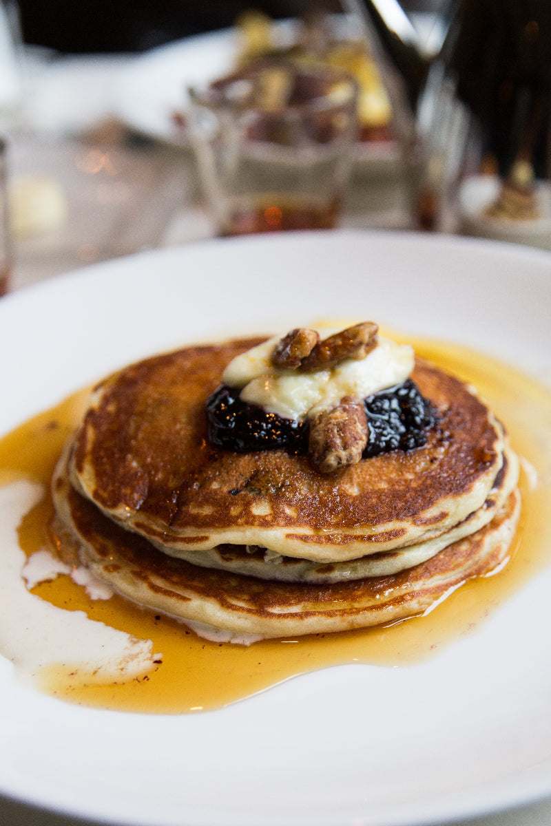 A stack of pancakes topped with banana slices, blackberries, and candied nuts, drizzled with syrup, served on a white plate.