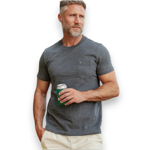 A man with a gray beard wearing the CRIQUET - SLUB COTTON SHORT SLEEVE POCKET T-SHIRT IN WASHED BLACK holds a canned drink in a green koozie outdoors, with trees and a white vehicle blurred in the background.