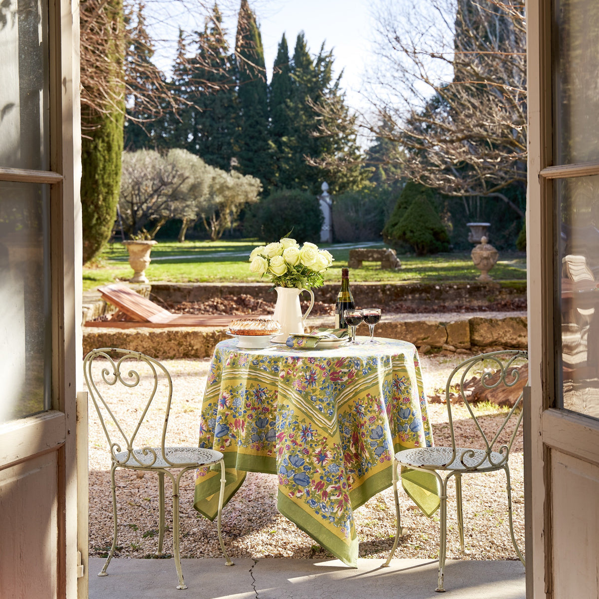 A sunlit outdoor dining scene showcases a COULEUR NATURE JARDIN BLEU &amp; VERT TABLECLOTH 59X59, set for two. A bouquet of white roses, a wine bottle, and glasses add elegance. The garden view, framed by open French doors, features trees and shrubs in the background.