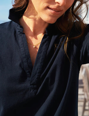 A woman in the FRANK & EILEEN EMMA Popover Henley Maxi Dress in British Royal Navy and sandals walks on a wooden deck beside an outdoor dining table, with a yellow straw bag and blanket nearby, and a house with large windows in the background.
