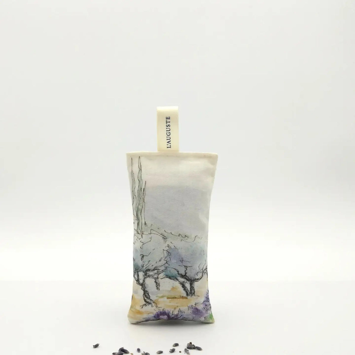 A rectangular L’AUGUSTE PROVENCE Lavender Sachet from Provence, featuring a scenic design and labeled fabric loop, stands upright on a white background with scattered Provence lavender buds in front.
