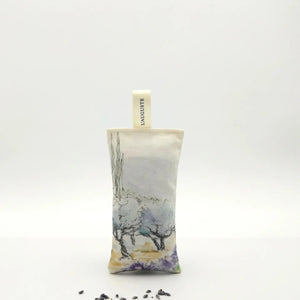 A rectangular L’AUGUSTE PROVENCE Lavender Sachet from Provence, featuring a scenic design and labeled fabric loop, stands upright on a white background with scattered Provence lavender buds in front.