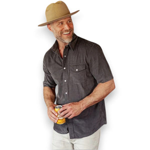 Close-up of a person wearing the CRIQUET Short Sleeve Corduroy Pearl Snap Shirt in Iron Horse, featuring pearl snap buttons and a chest pocket. Part of the person’s neck and chest is visible above the collar.