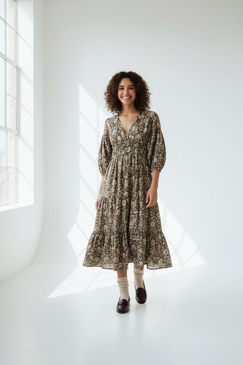 A woman stands against a gray background wearing the HOPE FOR FLOWERS TIERED PEASANT DRESS with puffed sleeves and a shirred bodice. She has her hands in its pockets and pairs dark loafers with light-colored socks.