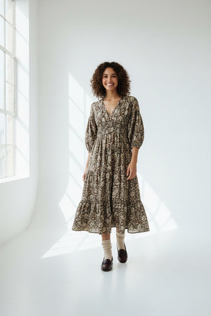 A woman stands against a gray background wearing the HOPE FOR FLOWERS TIERED PEASANT DRESS with puffed sleeves and a shirred bodice. She has her hands in its pockets and pairs dark loafers with light-colored socks.