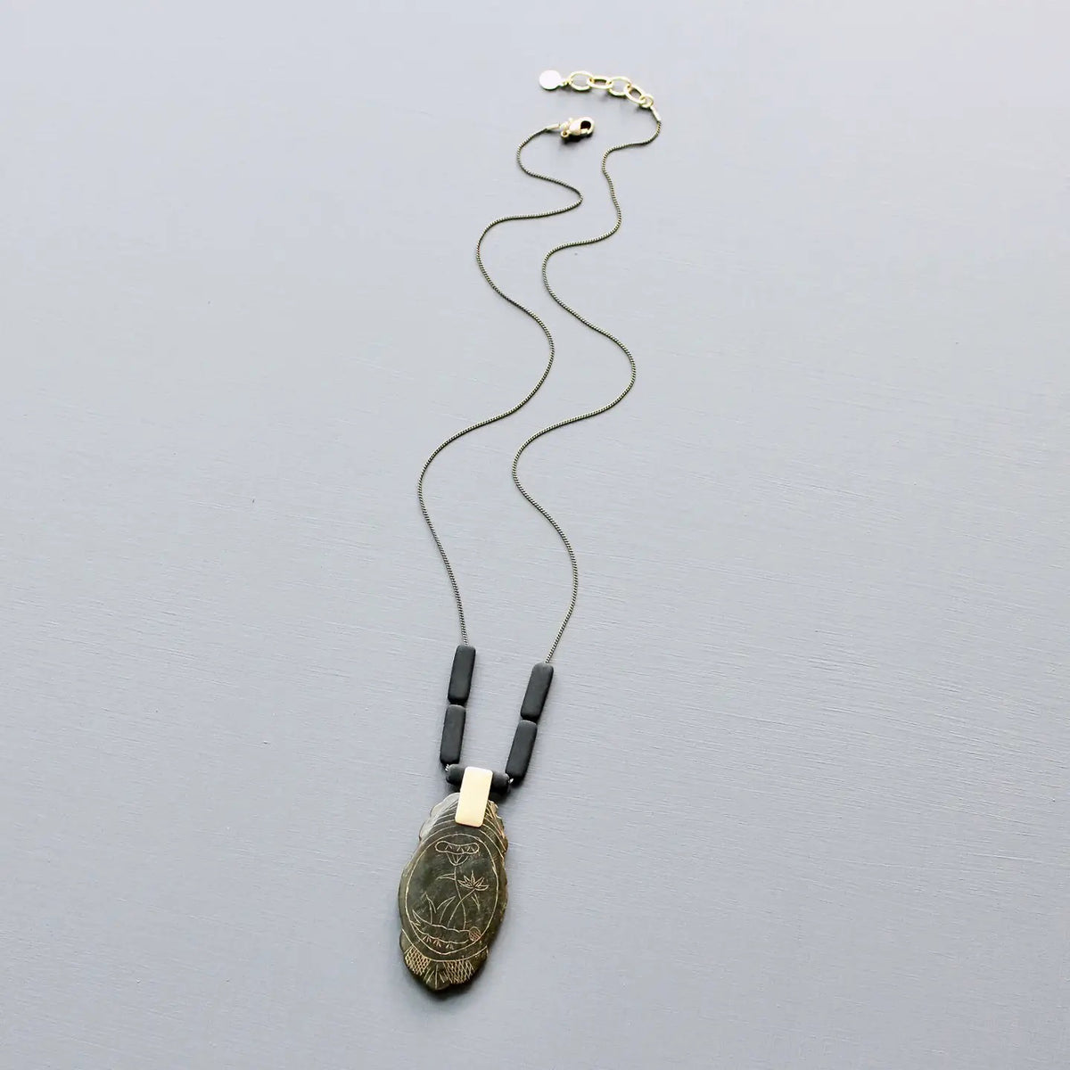 The DAVID AUBREY - CARVED JADE PENDANT NECKLACE by DAVID AUBREY features a 24-inch brass-ox rectangular chain with three black beads and a feather-shaped pendant engraved with an owl and plant design, displayed on a light grey background.